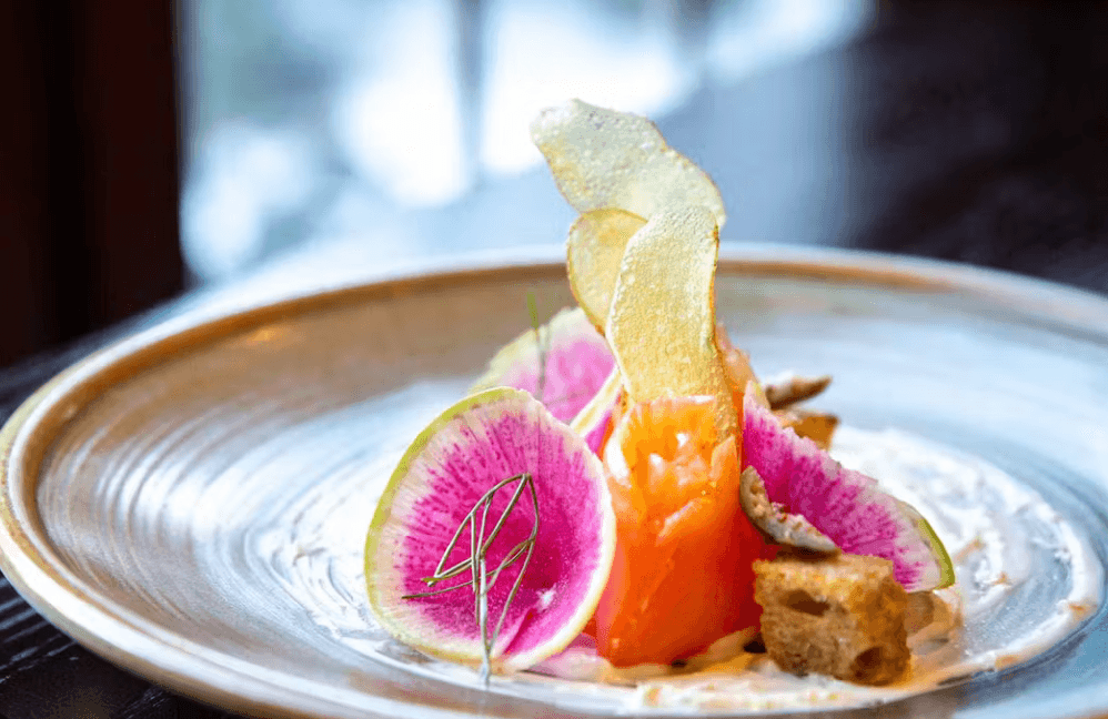 A gourmet dish inspired by the Culinary Institute, featuring thinly sliced watermelon radish, cured salmon, crispy chips, and garnishes, artfully arranged on a ceramic plate with a creamy sauce. The background is softly blurred.